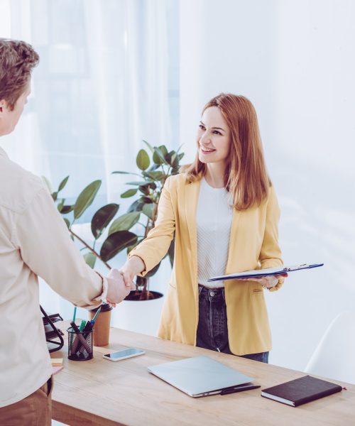 happy-recruiter-holding-clipboard-and-shaking-hands-with-employee-in-modern-office.jpg